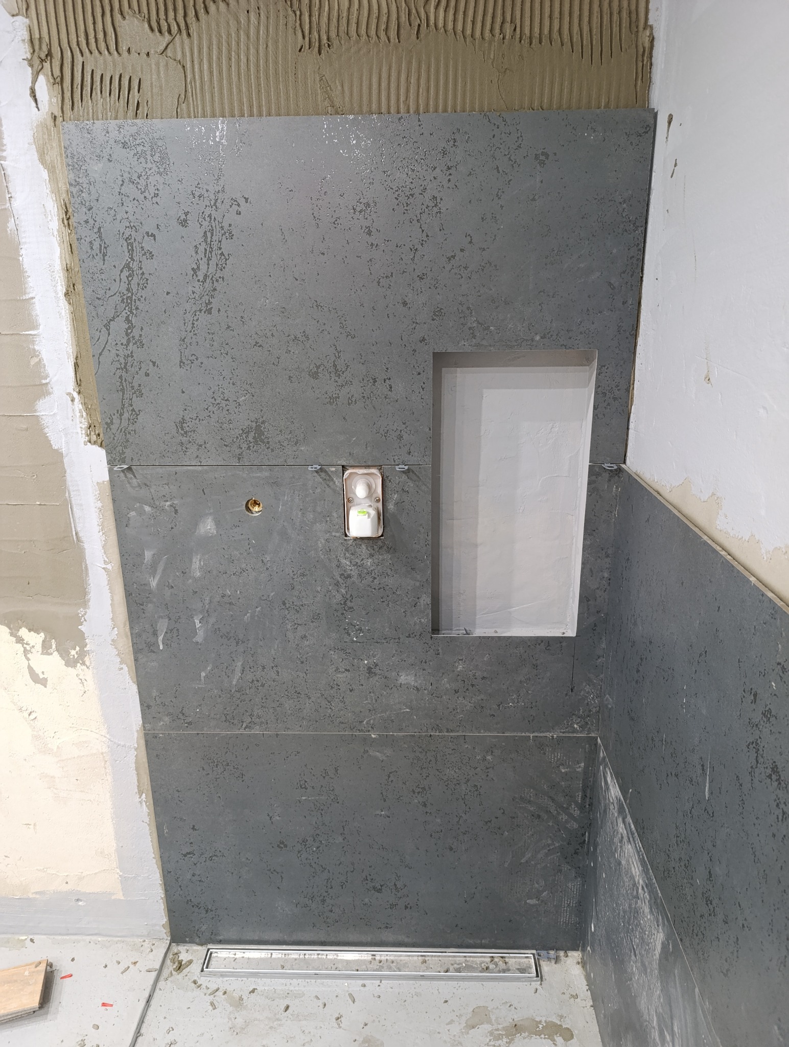 Montaż płytek imitujących kamień w łazience, widoczna wnęka w ścianie, odpływ liniowy w podłodze i fragment ściany przygotowanej pod płytki.