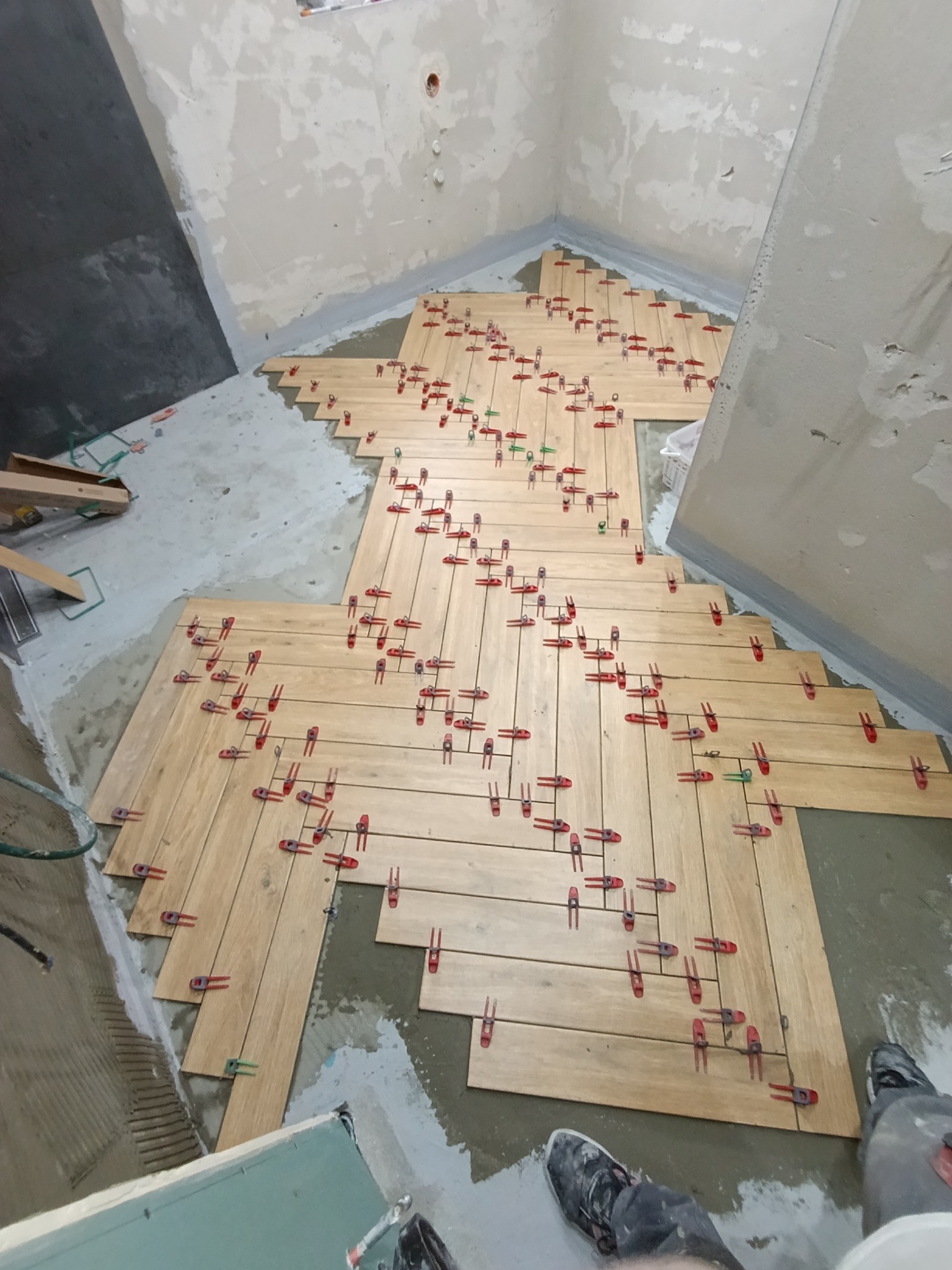 Układanie drewnopodobnych płytek w jodełkę w łazience, z użyciem czerwonych klipsów poziomujących. Widoczne niedokończone ściany i fragment butów osoby wykonującej prace.