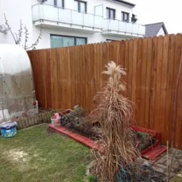 Nowe drewniane ogrodzenie panelowe oddziela ogród z małą szklarnią i rabatą od nowoczesnego domu z balkonami.