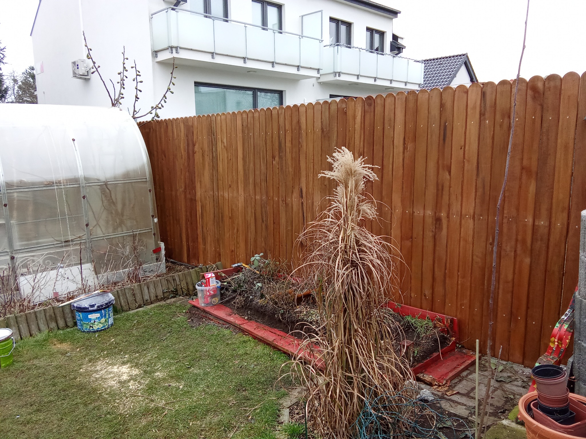 Nowe drewniane ogrodzenie panelowe oddziela ogród z małą szklarnią i rabatą od nowoczesnego domu z balkonami.