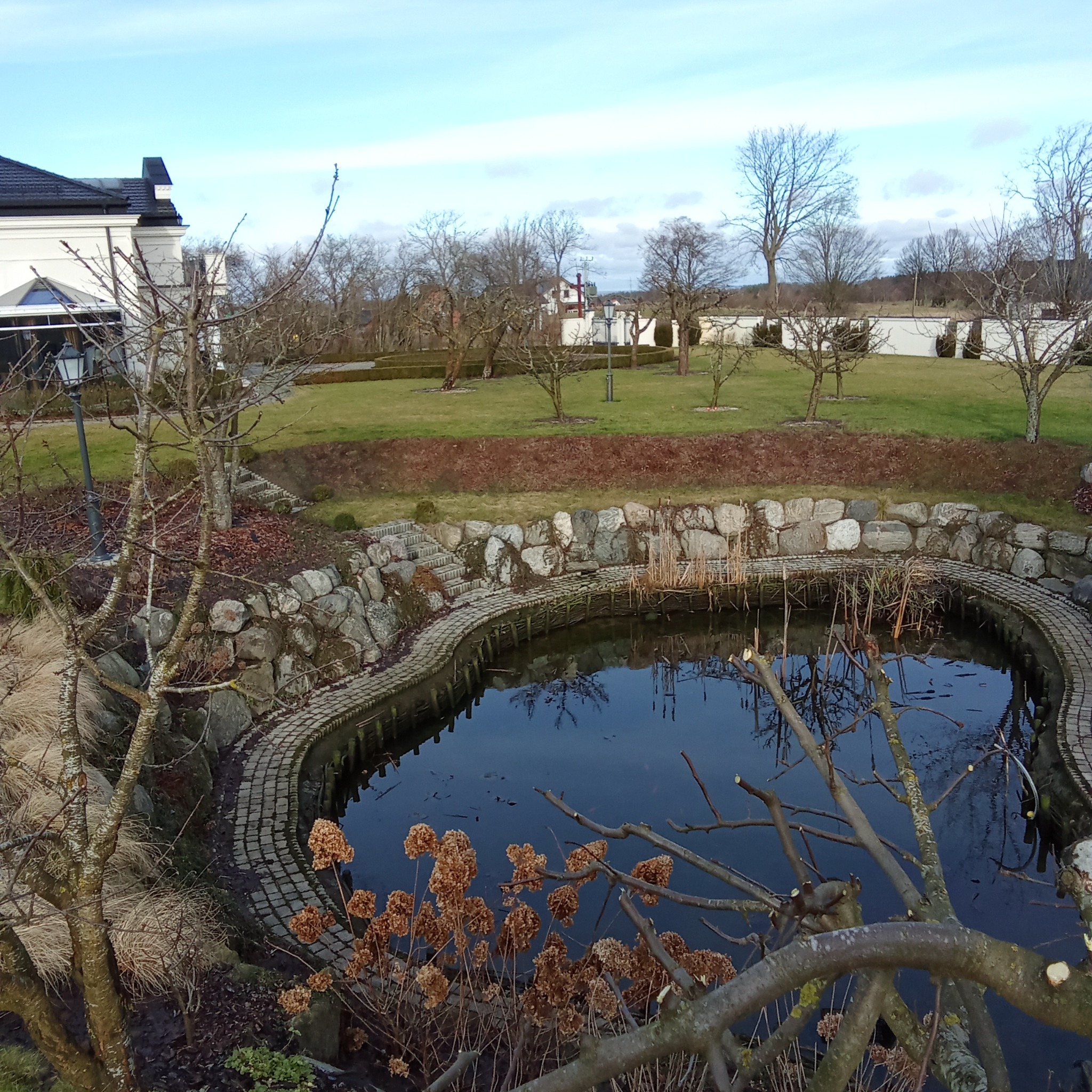 Oczko wodne z kamiennym brzegiem i ścieżką z kostki, widok z góry, w tle trawnik, drzewa i budynek.