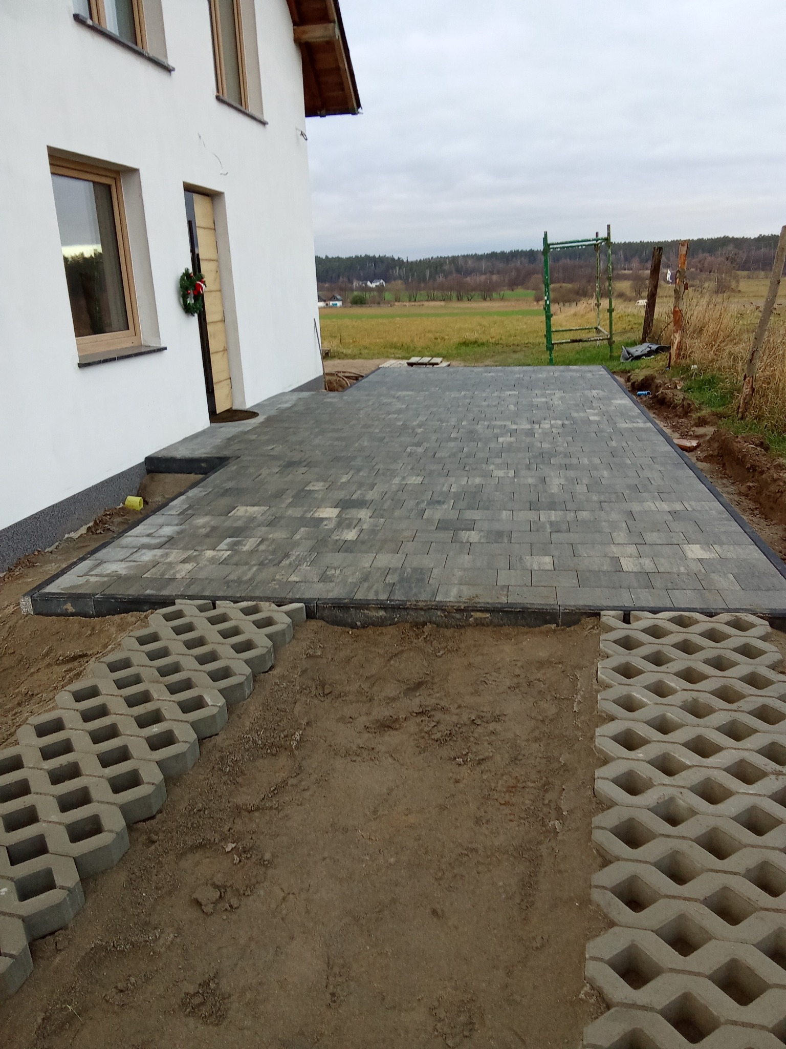 Ułożona szara kostka brukowa przed białym domem z wieńcem na drzwiach, widoczne elementy trawnika i krajobrazu w tle.