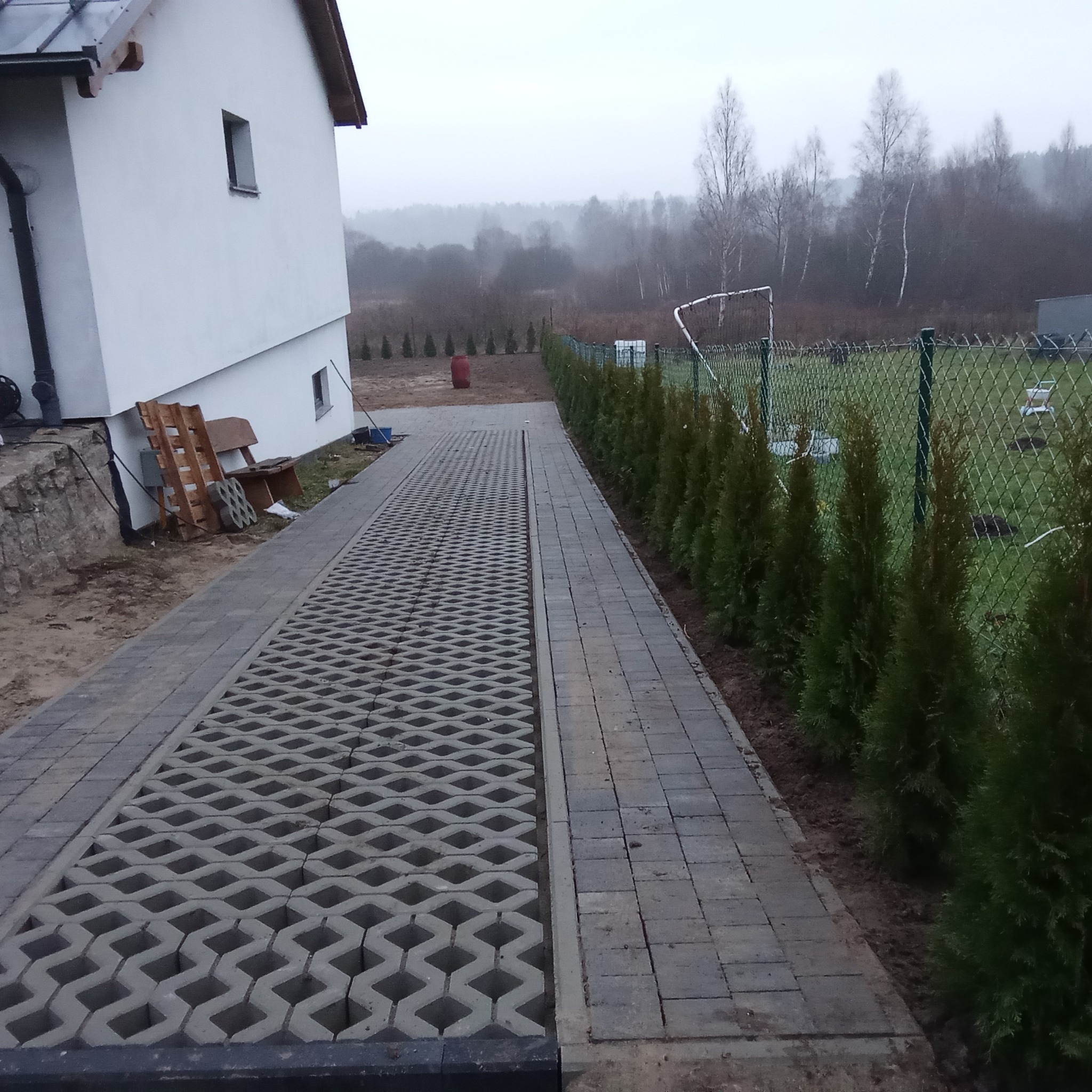 Ułożona ścieżka z kostki brukowej i ażurowej obok domu, z nasadzeniami iglaków przy ogrodzeniu z siatki.