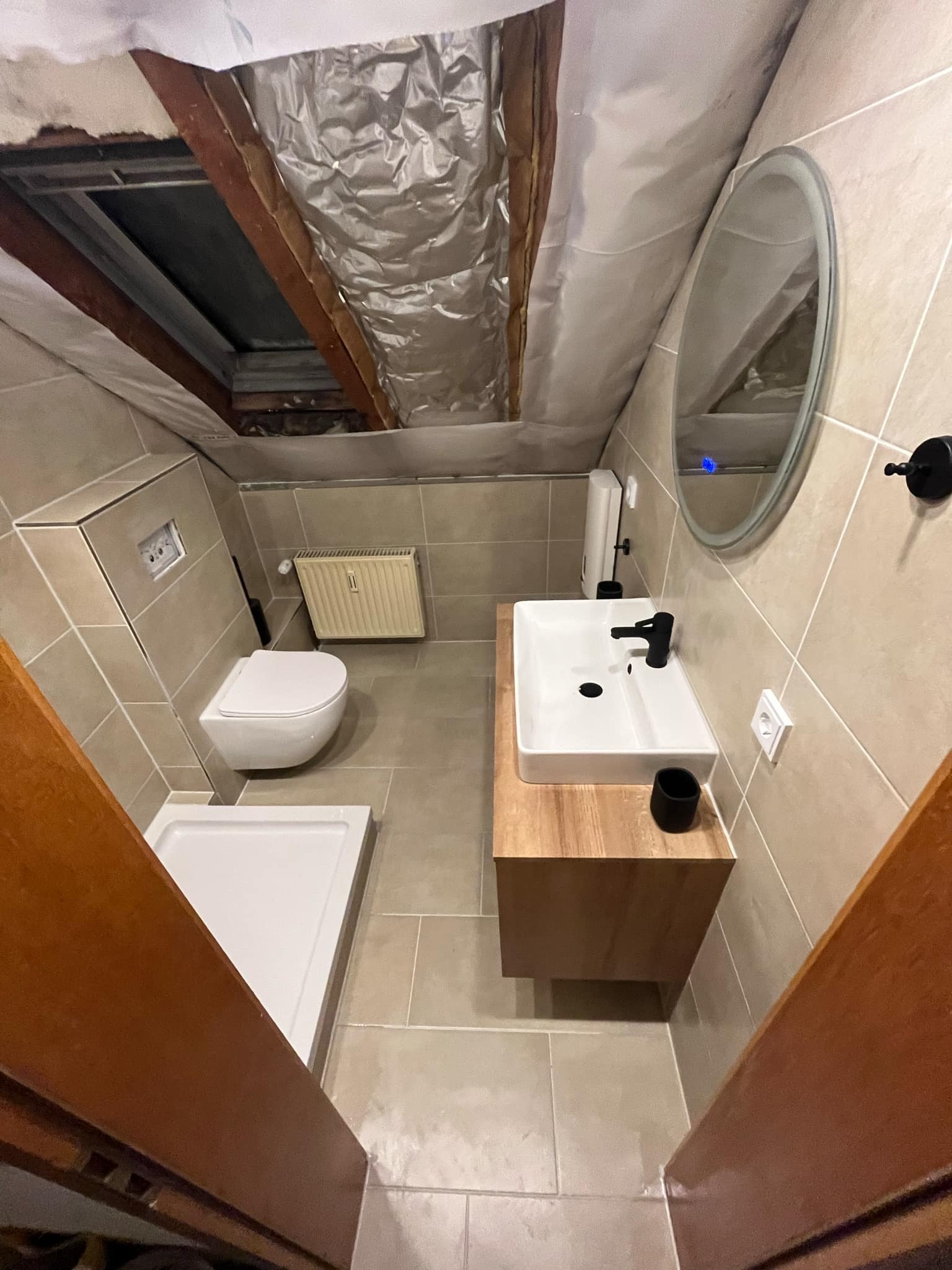 Ukończona łazienka na poddaszu ze skośnym sufitem, beżowe płytki, biała umywalka z czarną armaturą, toaleta podwieszana i brodzik.