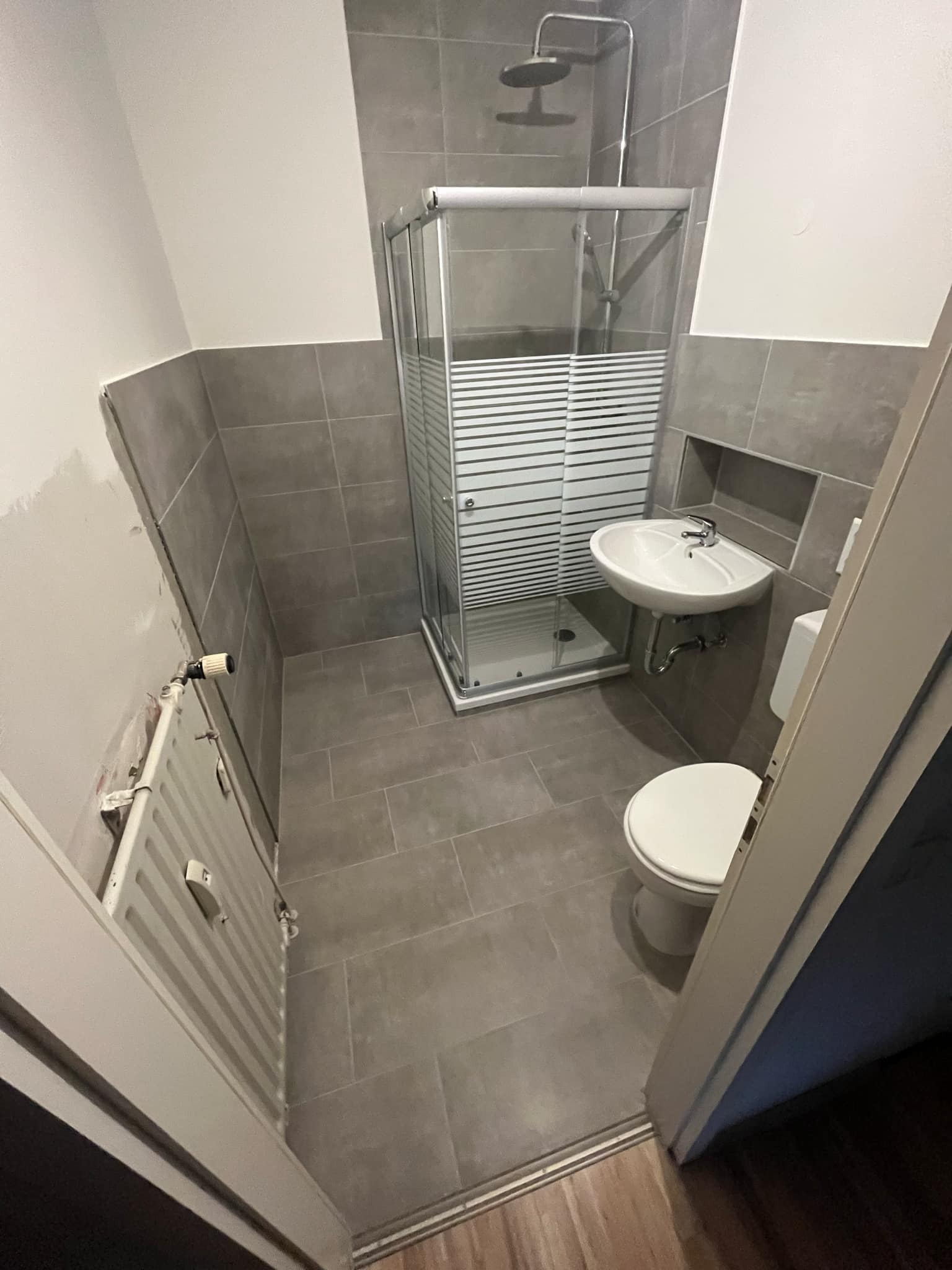 Szara łazienka z kabiną prysznicową z poziomymi paskami, białą umywalką w niszy, toaletą i grzejnikiem żeberkowym z termostatem, częściowo wykończona ściana z widocznymi śladami po remoncie.