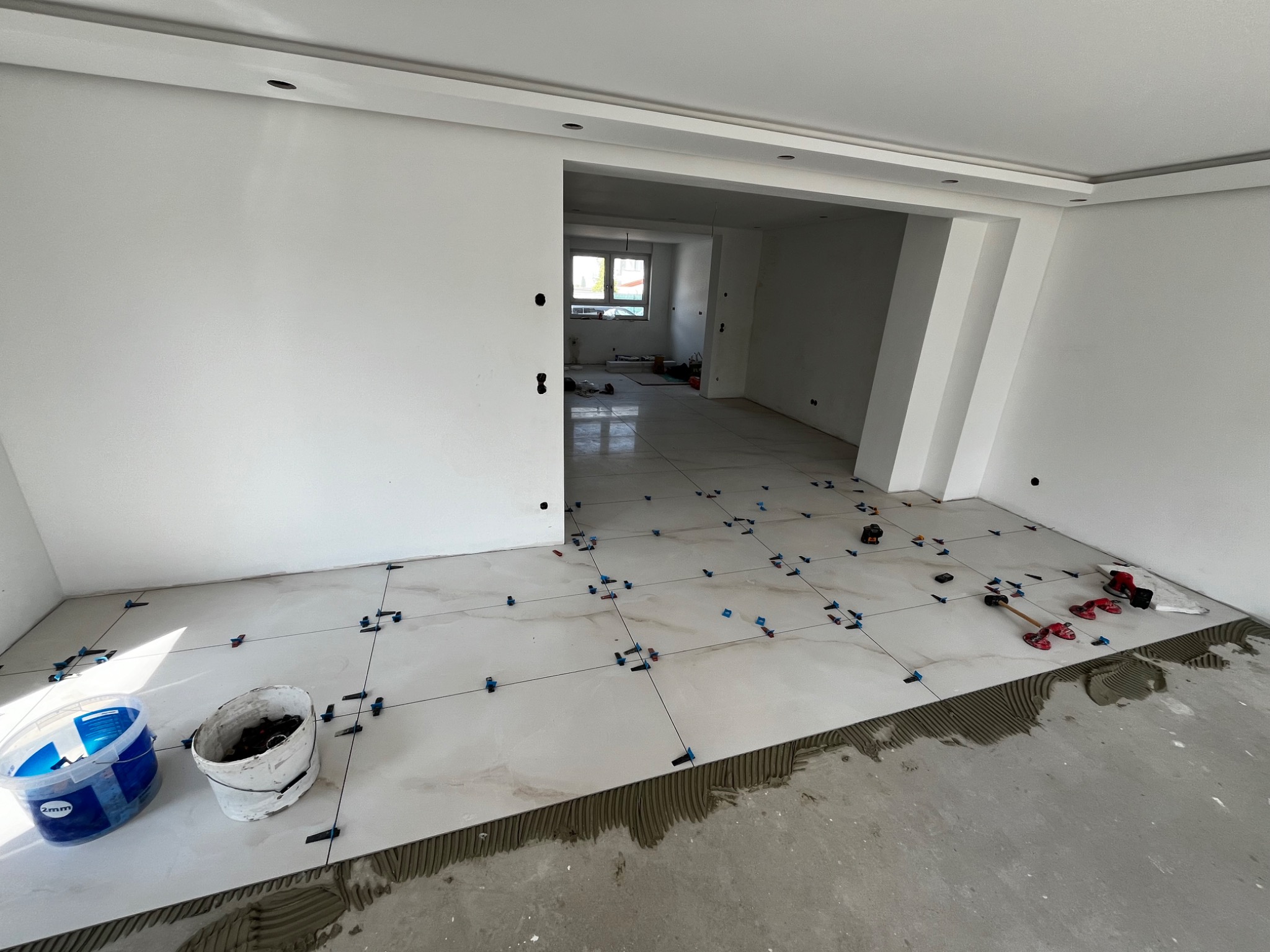 Układanie jasnych, marmurkowych płytek podłogowych w przestronnym, jasnym wnętrzu. Widoczne narzędzia glazurnicze, system poziomowania płytek oraz wiadro z akcesoriami.