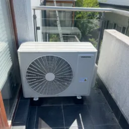 Biała jednostka zewnętrzna klimatyzacji Innova, zamontowana na czarnych podstawkach na balkonie z szarą balustradą i widokiem na zieleń.