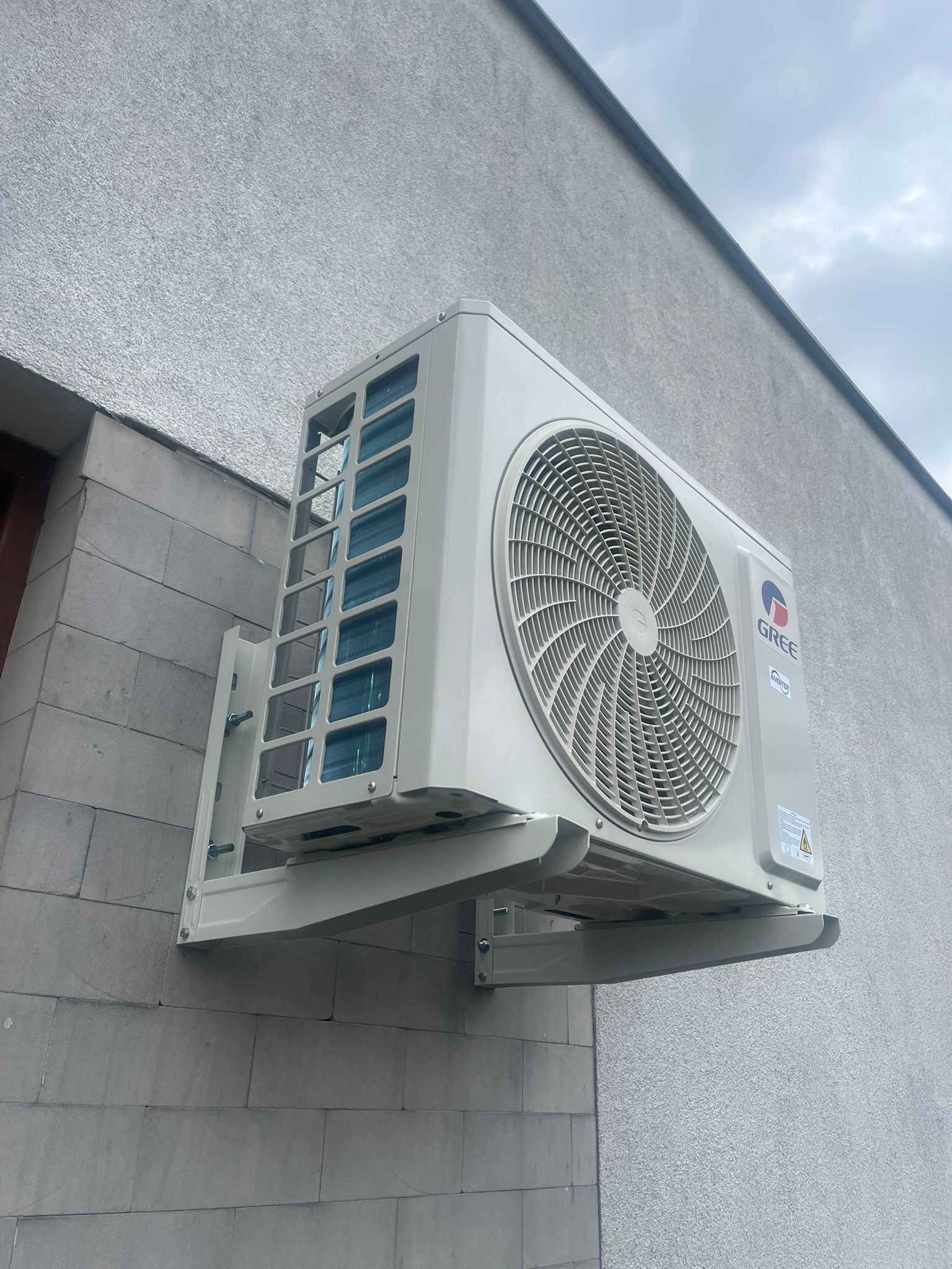 Zewnętrzna jednostka klimatyzacji marki Gree, zamontowana na szarej, teksturowanej ścianie budynku, widoczna z dołu pod kątem.