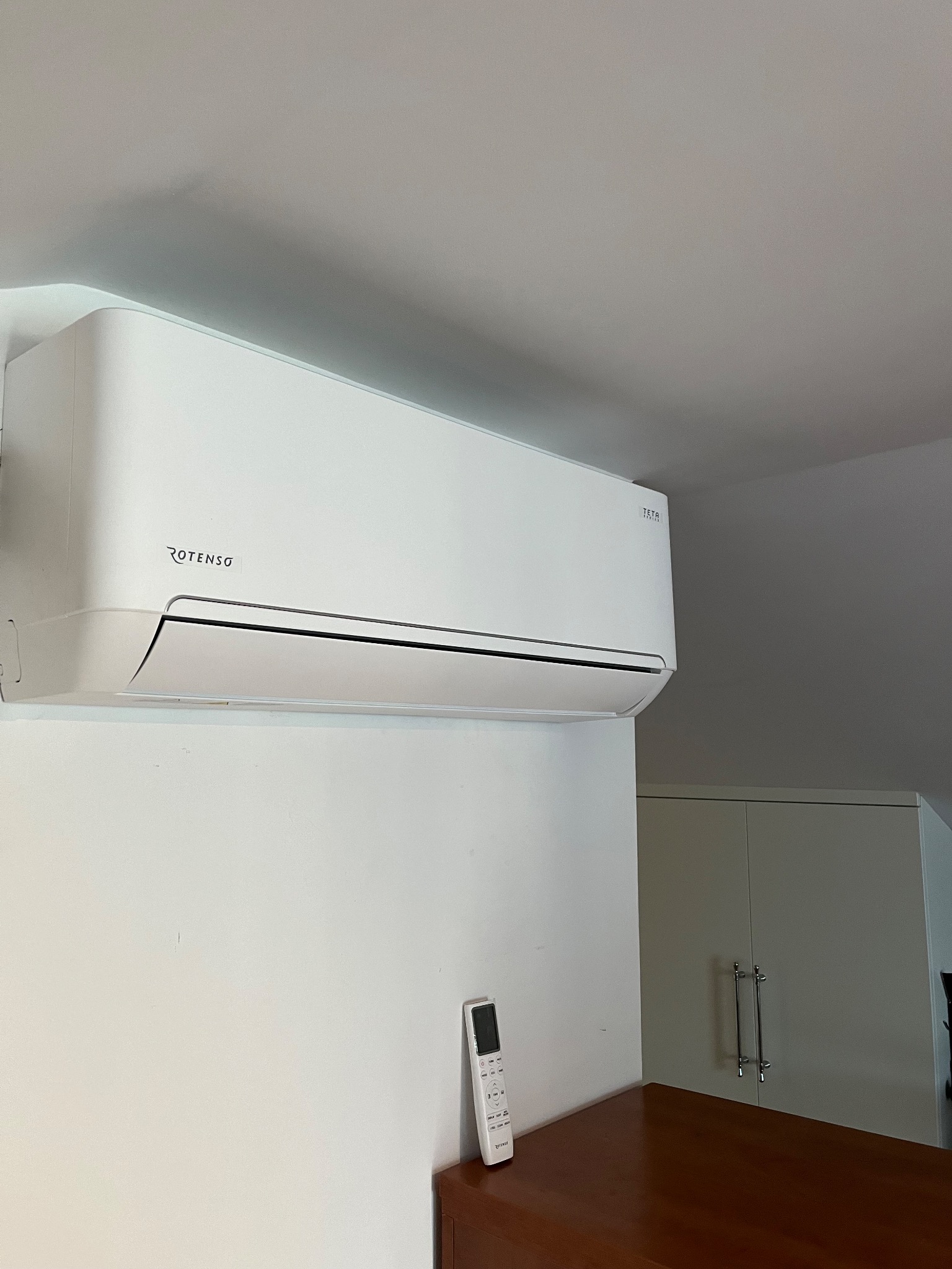 Biała klimatyzacja Rotenso TETA zamontowana na ścianie w pomieszczeniu z widocznym pilotem, szafką i sufitem.
