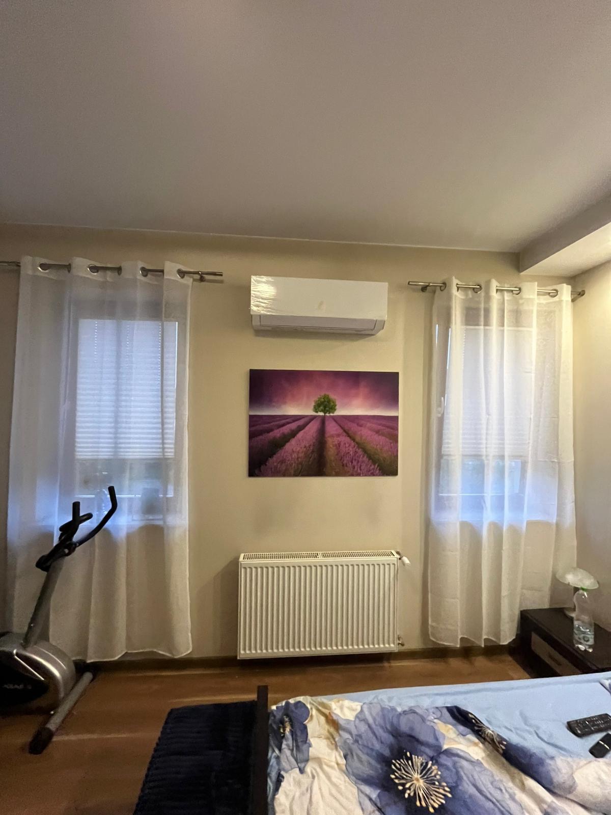 Klimatyzator ścienny w folii ochronnej, zamontowany w sypialni nad grzejnikiem i obrazem z motywem lawendy.