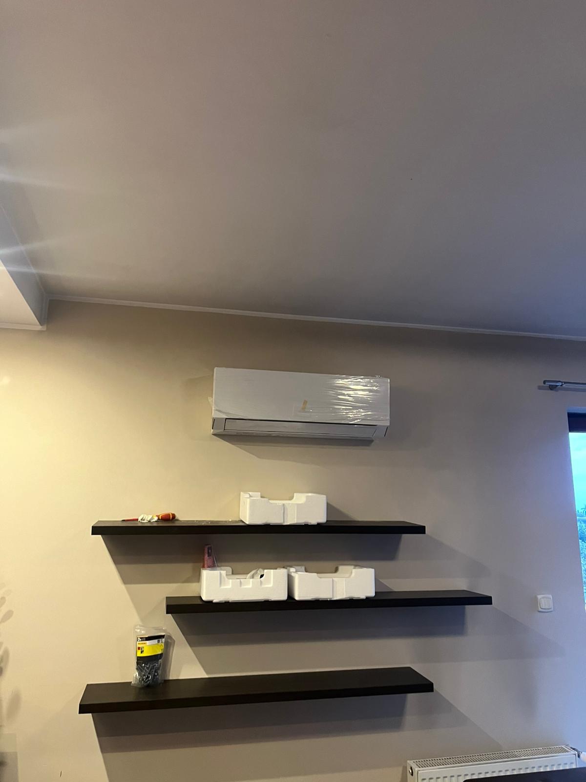 Zamontowany klimatyzator ścienny, jeszcze owinięty folią ochronną, widoczny na ścianie nad trzema czarnymi półkami, na których znajdują się narzędzia i elementy montażowe.