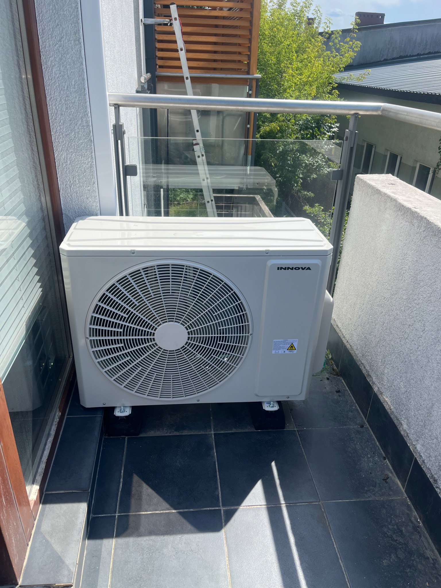 Biała jednostka zewnętrzna klimatyzacji Innova, zamontowana na czarnych podstawkach na balkonie z szarą balustradą i widokiem na zieleń.