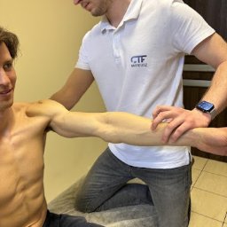 Centrum Treningu i Fizjoterapii Bieruń - Fizjoterapia - Masaż - Trening - Mężczyzna w trakcie badania zakresu ruchu ramienia przez terapeutę w białej koszulce polo z logo firmy, pacjent uśmiechnięty.