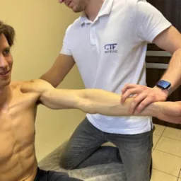 Mężczyzna w trakcie badania zakresu ruchu ramienia przez terapeutę w białej koszulce polo z logo firmy, pacjent uśmiechnięty.