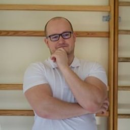 Centrum Treningu i Fizjoterapii Bieruń - Fizjoterapia - Masaż - Trening - Mężczyzna w okularach i białej koszulce, z ręką pod brodą, stoi na tle drabinek gimnastycznych w jasnym pomieszczeniu.