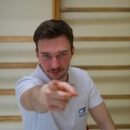 Centrum Treningu i Fizjoterapii Bieruń - Fizjoterapia - Masaż - Trening - Młody mężczyzna w białej koszulce z logo, wskazujący palcem w kierunku widza na tle ściany z drabinką gimnastyczną.