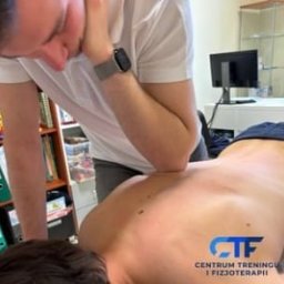 Centrum Treningu i Fizjoterapii Bieruń - Fizjoterapia - Masaż - Trening - Mężczyzna w białej koszulce, z zegarkiem na lewym nadgarstku, wykonuje masaż łokciem na plecach pacjenta leżącego na stole do masażu. W tle widoczny regał z książkami i segregatorami oraz monitor...