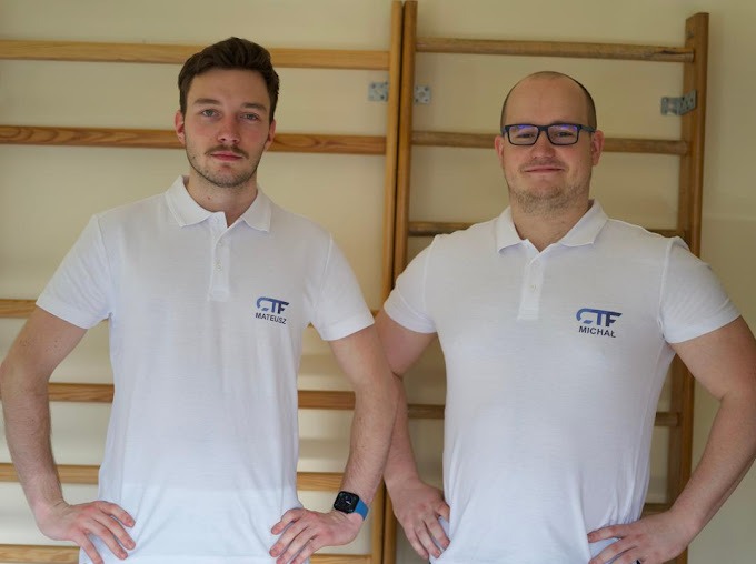 Dwóch mężczyzn w białych koszulkach polo z logo firmy, stojących na tle drabinek gimnastycznych.