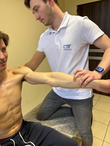 Mężczyzna w trakcie badania zakresu ruchu ramienia przez terapeutę w białej koszulce polo z logo firmy, pacjent uśmiechnięty.