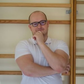 Mężczyzna w okularach i białej koszulce, z ręką pod brodą, stoi na tle drabinek gimnastycznych w jasnym pomieszczeniu.