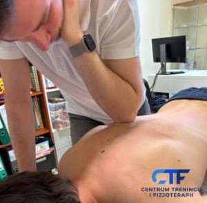 Mężczyzna w białej koszulce, z zegarkiem na lewym nadgarstku, wykonuje masaż łokciem na plecach pacjenta leżącego na stole do masażu. W tle widoczny regał z książkami i segregatorami oraz monitor...