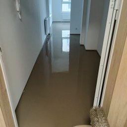 PatPanele Patryk Ninard - Świeżo wylana, gładka, samopoziomująca posadzka w korytarzu mieszkania, widok zza progu, z widocznym wiadrem i wałkiem kolczastym do odpowietrzania w dolnej części kadru.