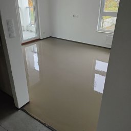 PatPanele Patryk Ninard - Świeżo wylana, gładka, samopoziomująca posadzka w pustym, jasnym pomieszczeniu z oknem i wyjściem na balkon; widoczne ślady po odpowietrzaniu.