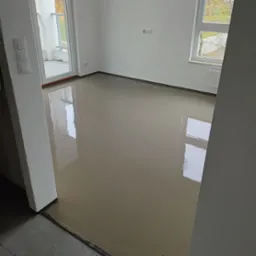 Świeżo wylana, gładka, samopoziomująca posadzka w pustym, jasnym pomieszczeniu z oknem i wyjściem na balkon; widoczne ślady po odpowietrzaniu.