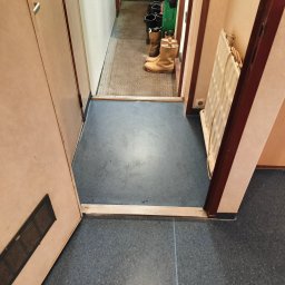 PatPanele Patryk Ninard - Szara, nakrapiana podłoga z widocznymi śladami użytkowania w korytarzu, z progiem i drzwiami po lewej oraz grzejnikiem po prawej stronie, w tle widoczne buty robocze.