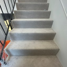 PatPanele Patryk Ninard - Schody wyłożone jasną, teksturowaną wykładziną dywanową. Metalowa barierka zabezpieczona folią. Widok z dołu na górę, podkreślający fakturę materiału.