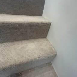 PatPanele Patryk Ninard - Beżowa wykładzina dywanowa na schodach, widok z bliska, delikatna tekstura, jasne oświetlenie, minimalistyczne tło ściany. Detal wykończenia wnętrza.