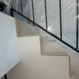 PatPanele Patryk Ninard - Schody z jasną wykładziną dywanową, zabezpieczone folią ochronną. Widoczne czarne metalowe barierki. Ujęcie z dołu, podkreślające detale wykończenia.