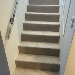 PatPanele Patryk Ninard - Schody wyłożone beżową wykładziną dywanową, poręcz zabezpieczona folią ochronną. Widok z dołu, jasne ściany, nowoczesne wykończenie.