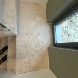 PatPanele Patryk Ninard - Wykończone schody z beżową wykładziną dywanową, widok z góry. Obok ściana z oknem w ciemnej ramie i fragment podłogi. Widoczne buty na schodach.