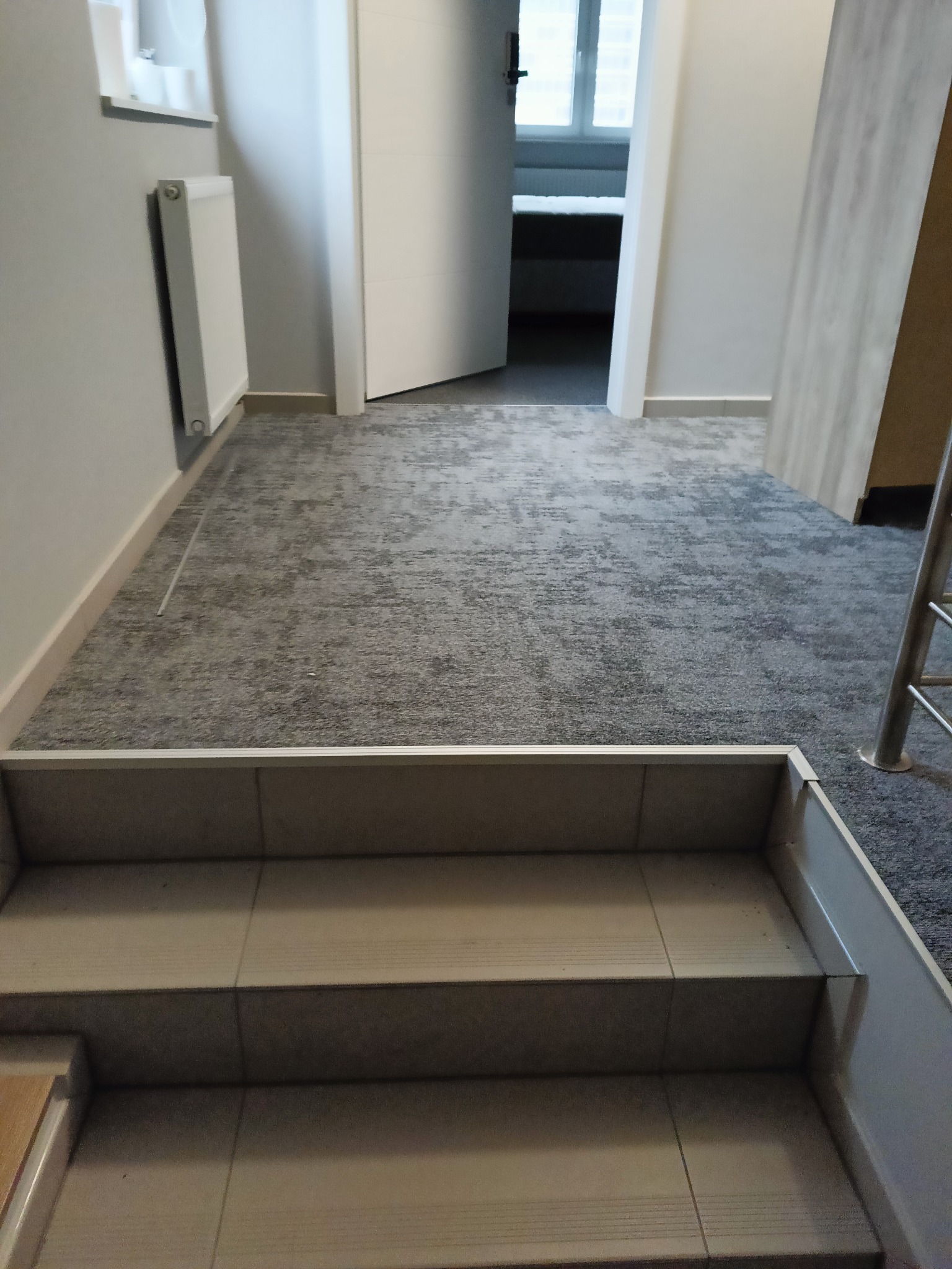 Szara wykładzina dywanowa z wzorem na podłodze w korytarzu, widok od strony schodów z ceramicznymi stopniami.