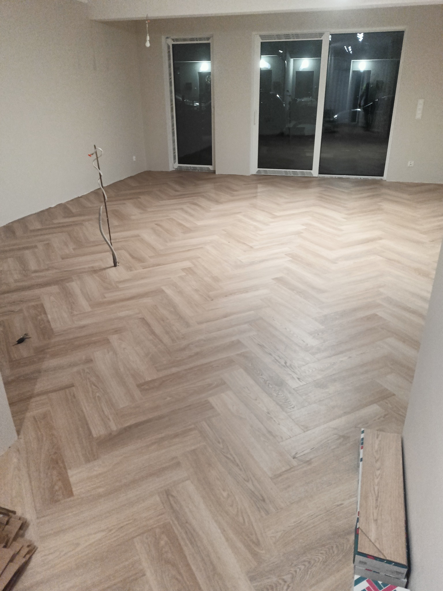 Ułożona podłoga z paneli w jodełkę w jasnym pomieszczeniu z dużymi oknami balkonowymi, widoczne wystające kable elektryczne i resztki paneli przy ścianie.