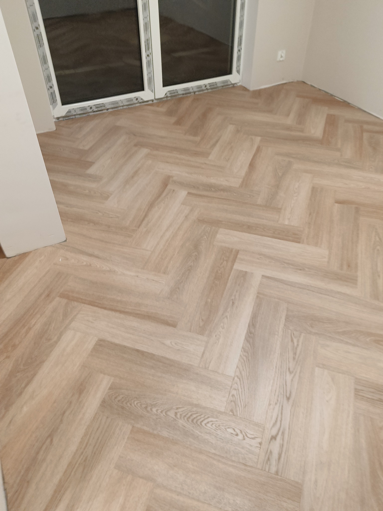 Jasne panele ułożone we wzór jodełki francuskiej w nowym pokoju, widok z góry, widoczne białe listwy przypodłogowe i okno.