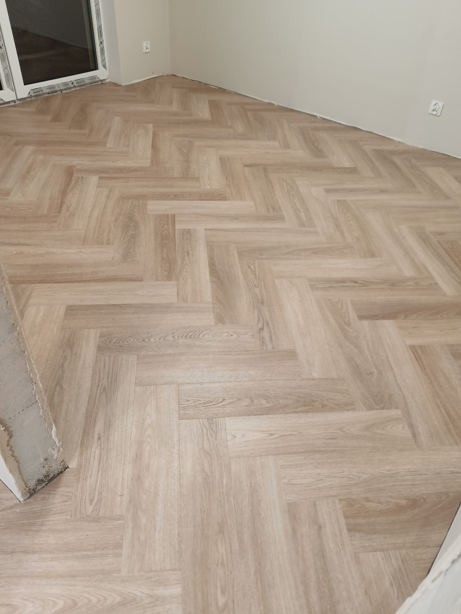 Ułożone panele podłogowe w jodełkę w jasnym odcieniu drewna, widoczny fragment ściany z gniazdkiem elektrycznym oraz fragment ściany przy drzwiach z widocznymi pozostałościami zaprawy.