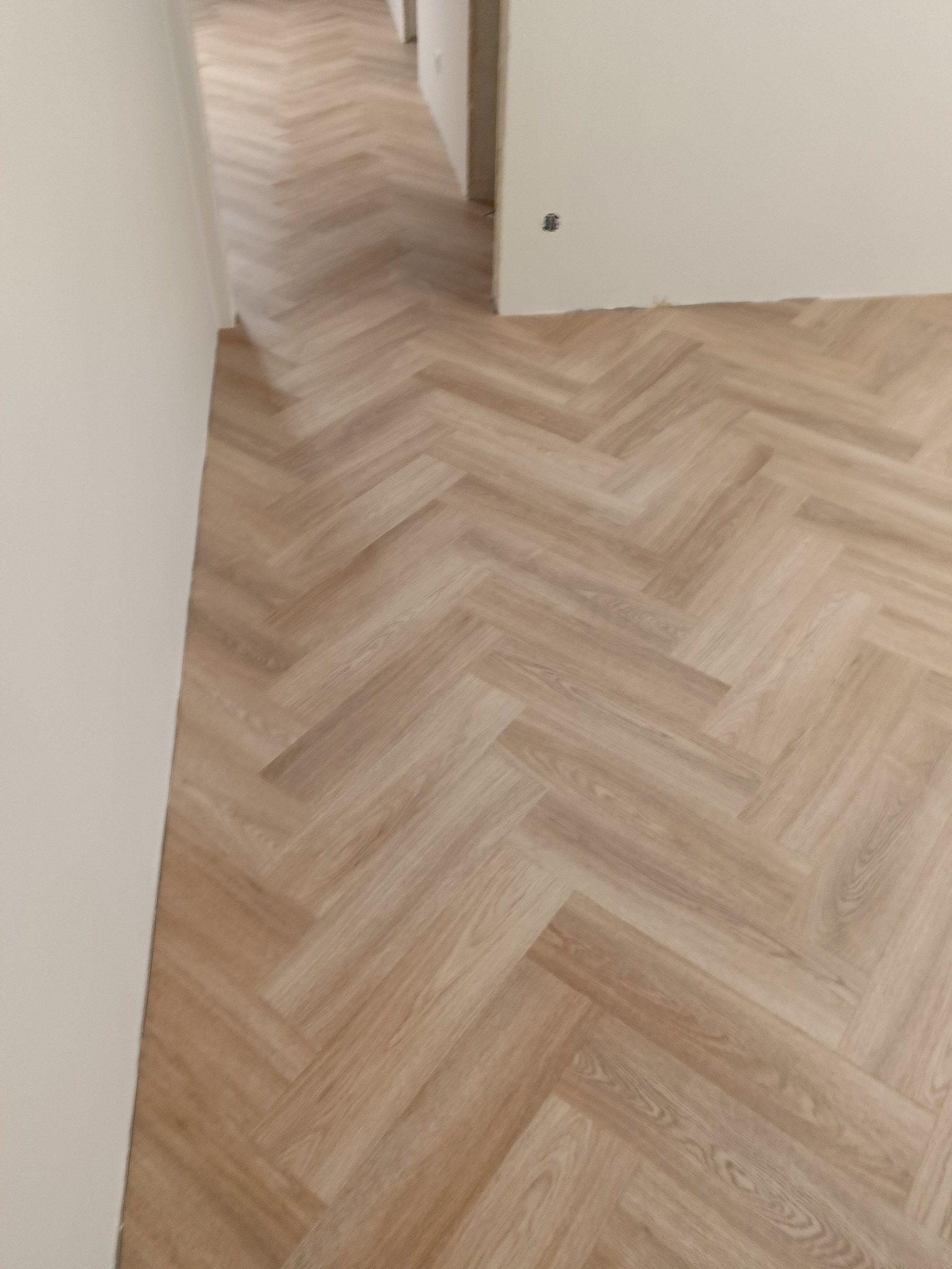 Jasne panele ułożone w jodełkę francuską, widok na korytarz w trakcie remontu, gniazdko elektryczne wystaje ze ściany.