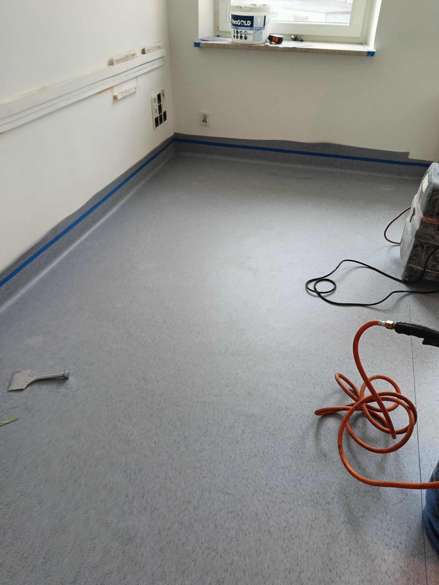 Szara podłoga z wykładziny PCV zabezpieczona niebieską taśmą malarską przy ścianach w Gdyni, widoczne narzędzia i maszyna do zgrzewania wykładzin.