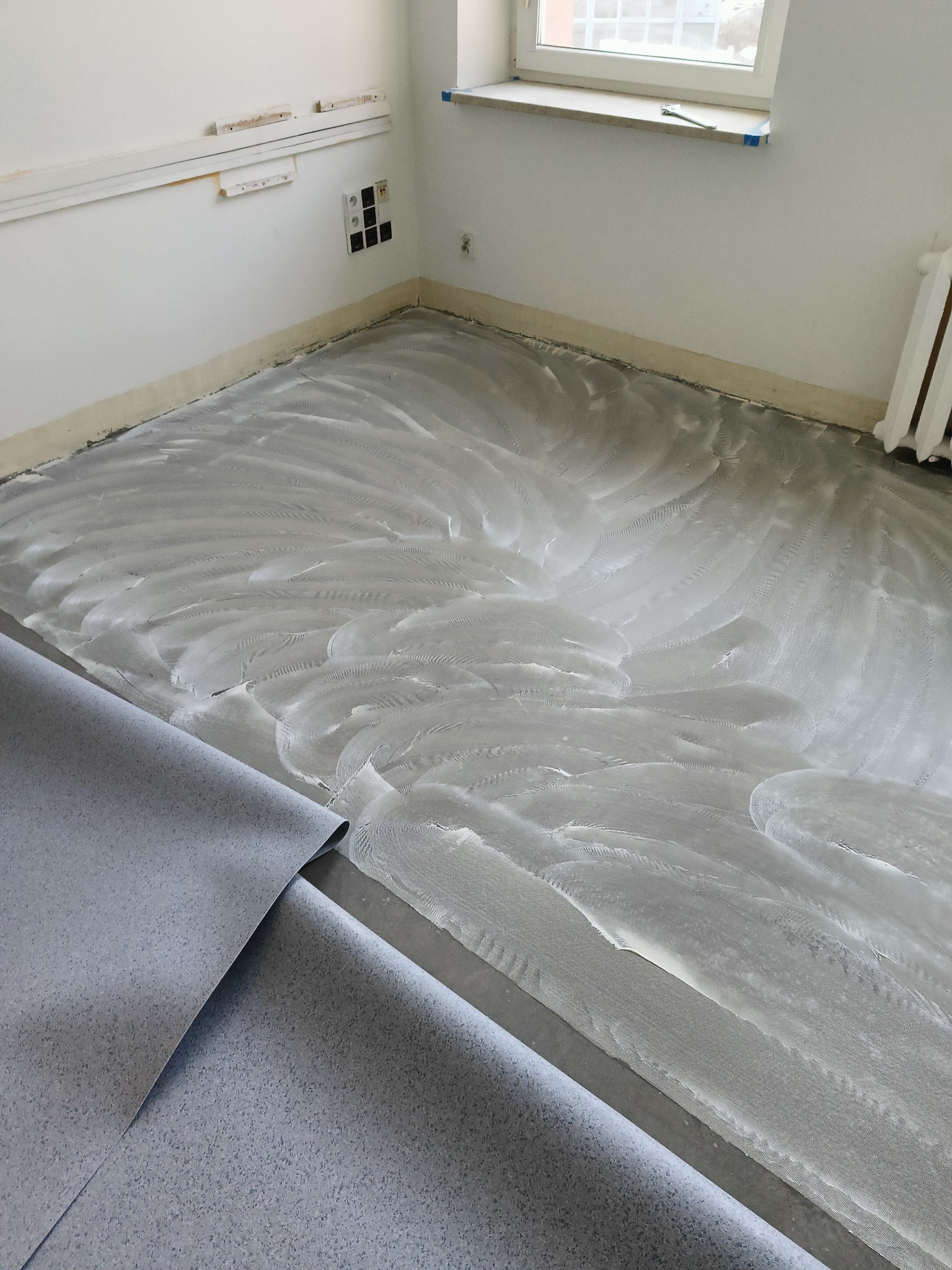 Przygotowanie podłoża pod nową podłogę: warstwa kleju z widocznymi śladami szpachli, częściowo rozłożony szary materiał podłogowy w rolce, białe ściany z widocznymi gniazdkami elektrycznymi.