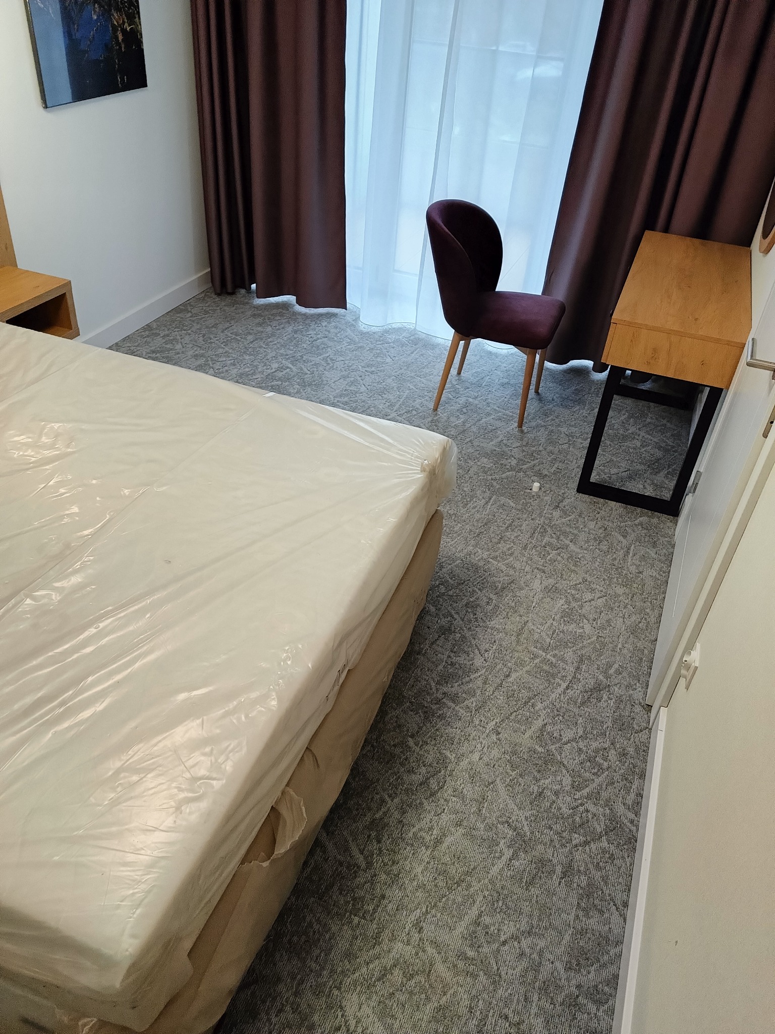 Pokój hotelowy z nową wykładziną dywanową w odcieniach szarości, zabezpieczone łóżko folią ochronną, widoczne brązowe zasłony i krzesło z ciemnej tkaniny.