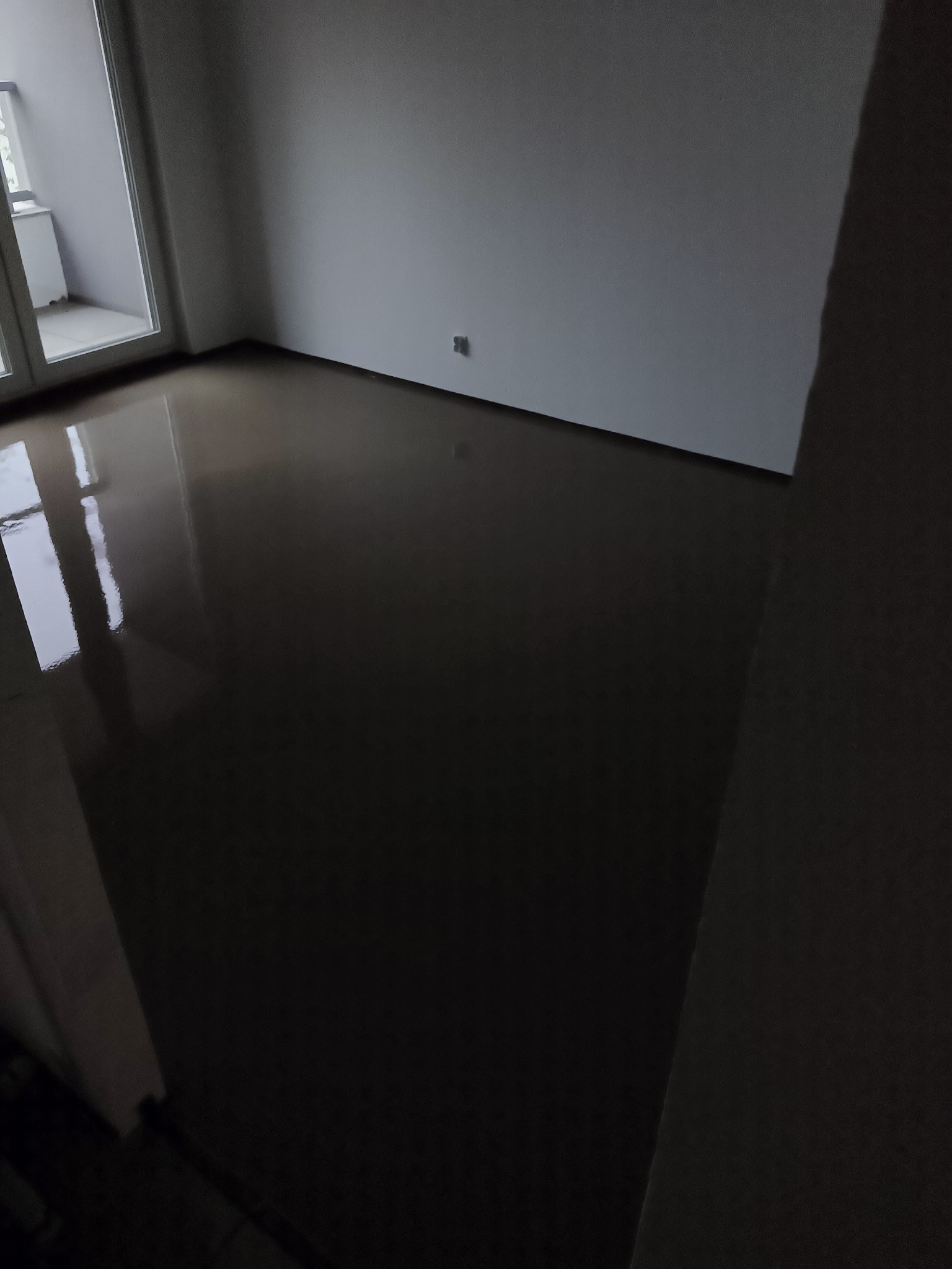 Świeżo wylana, ciemna posadzka w pomieszczeniu z balkonem, widoczne odbicia światła w gładkiej powierzchni.