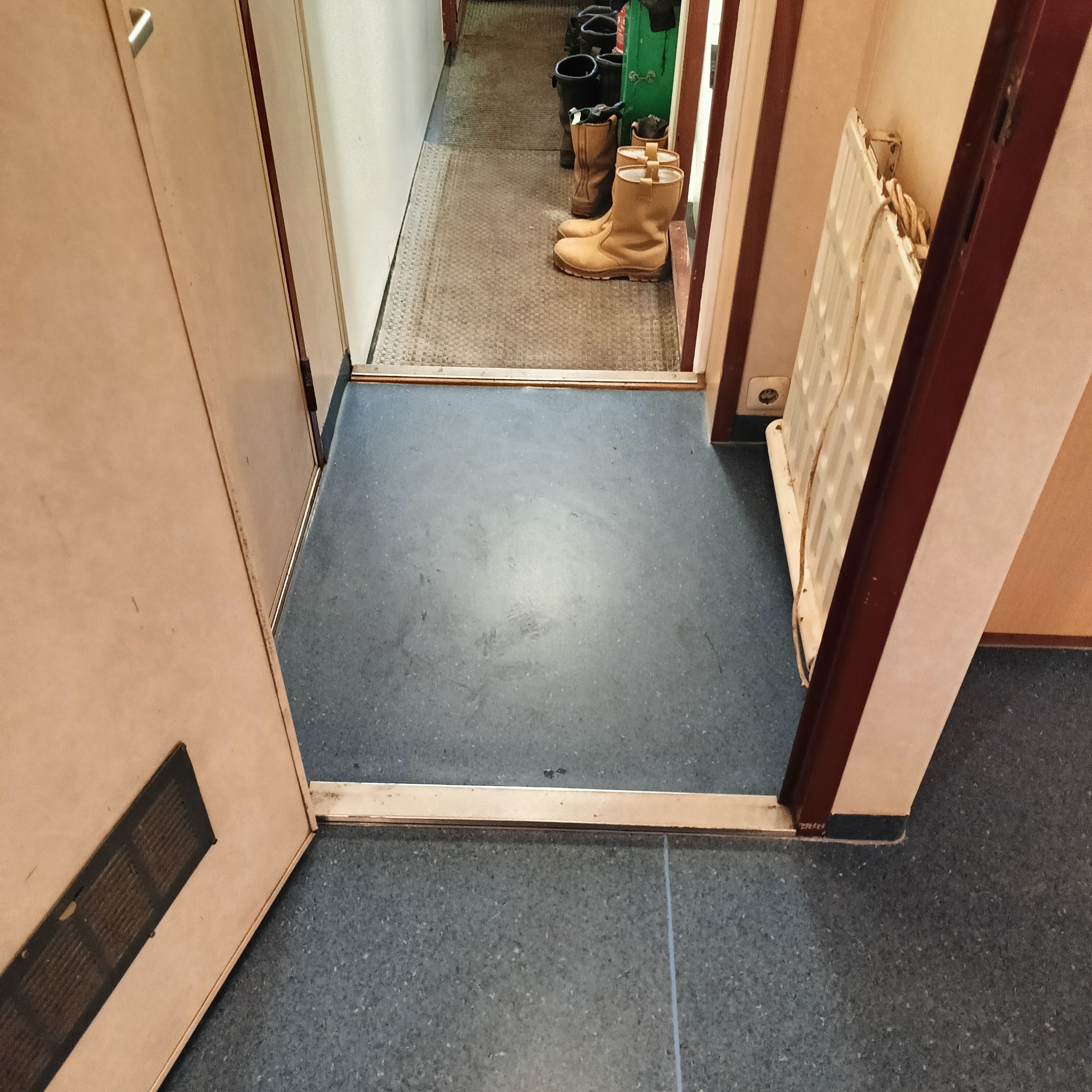 Szara, nakrapiana podłoga z widocznymi śladami użytkowania w korytarzu, z progiem i drzwiami po lewej oraz grzejnikiem po prawej stronie, w tle widoczne buty robocze.