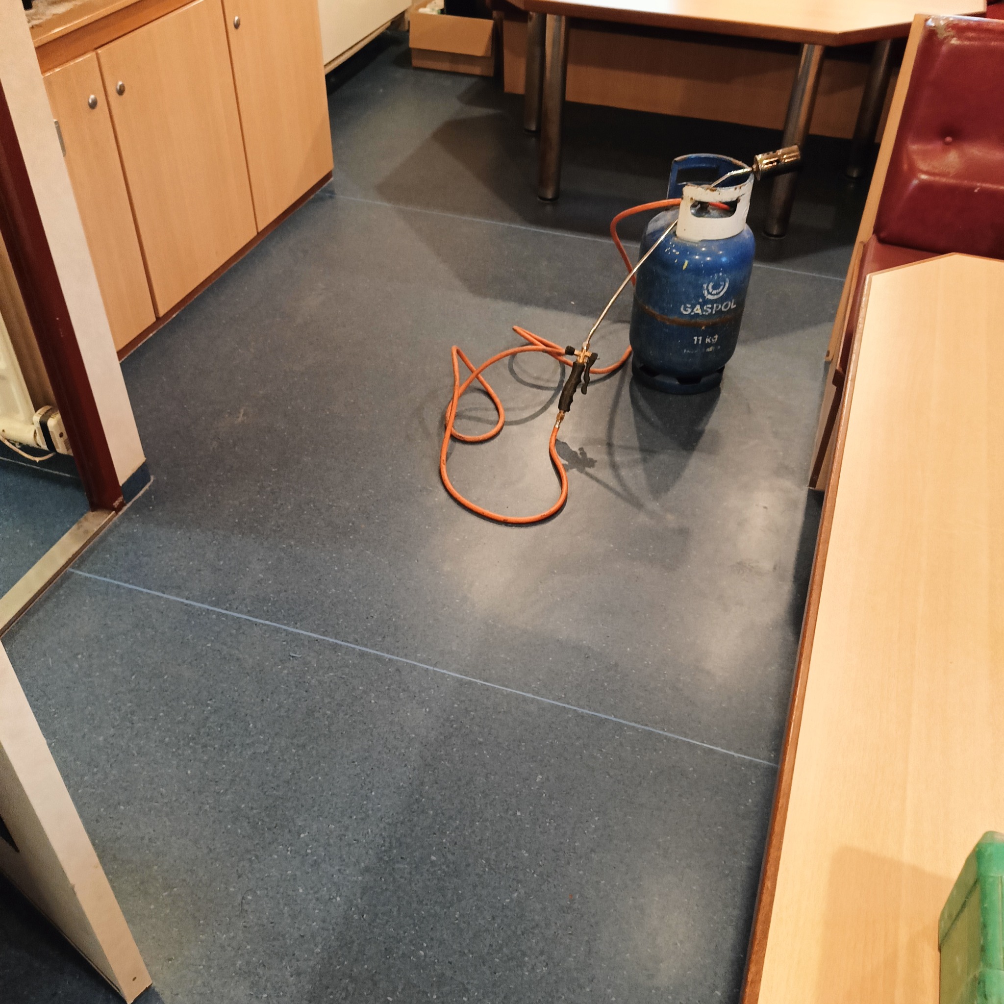 Niebieska butla gazowa z palnikiem i pomarańczowym przewodem leżąca na szarej posadzce w pomieszczeniu z meblami.