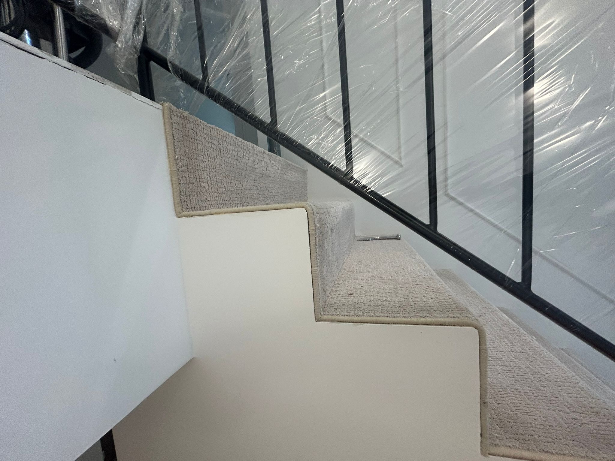 Schody z jasną wykładziną dywanową, zabezpieczone folią ochronną. Widoczne czarne metalowe barierki. Ujęcie z dołu, podkreślające detale wykończenia.