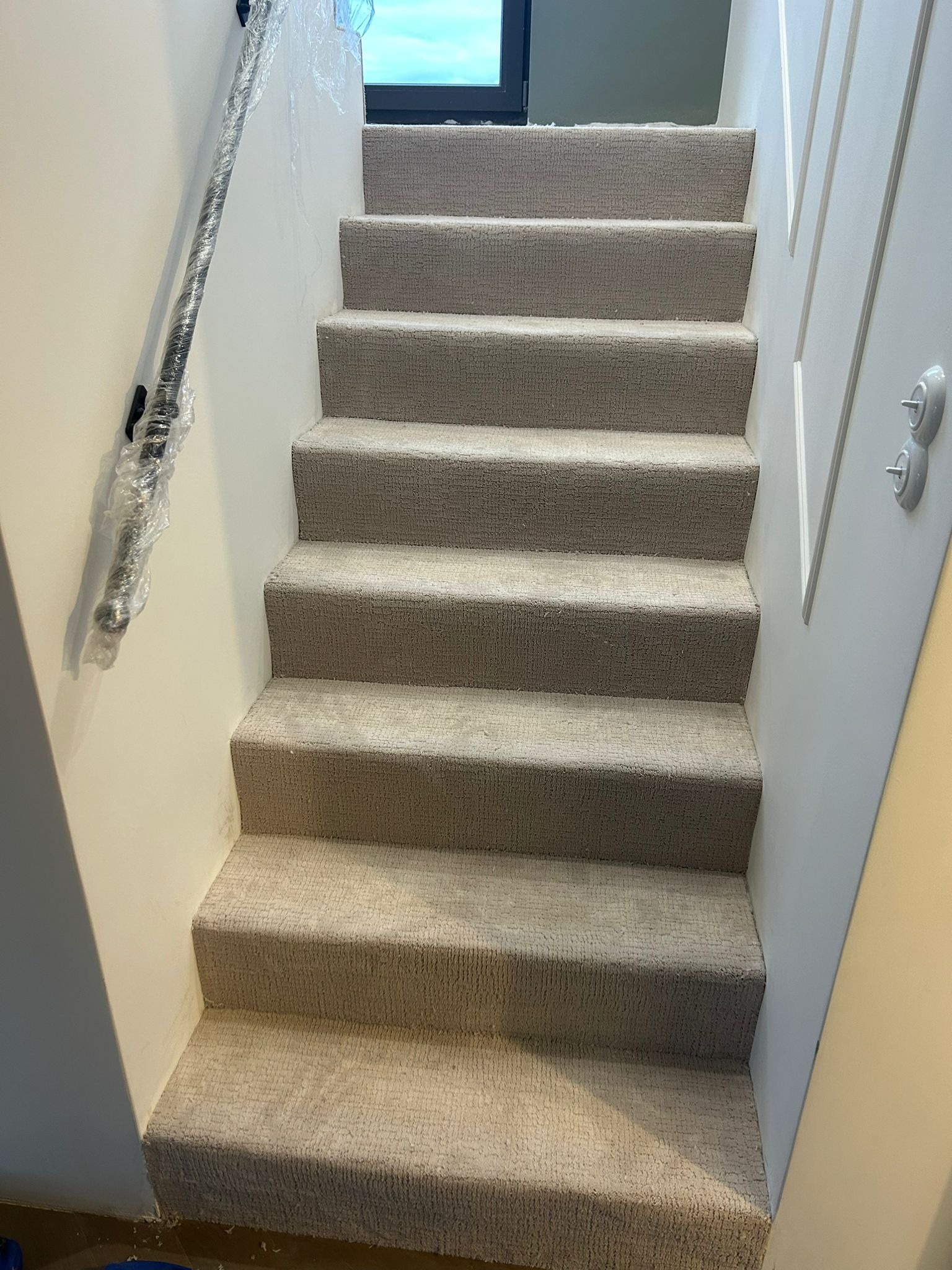 Schody wyłożone beżową wykładziną dywanową, poręcz zabezpieczona folią ochronną. Widok z dołu, jasne ściany, nowoczesne wykończenie.
