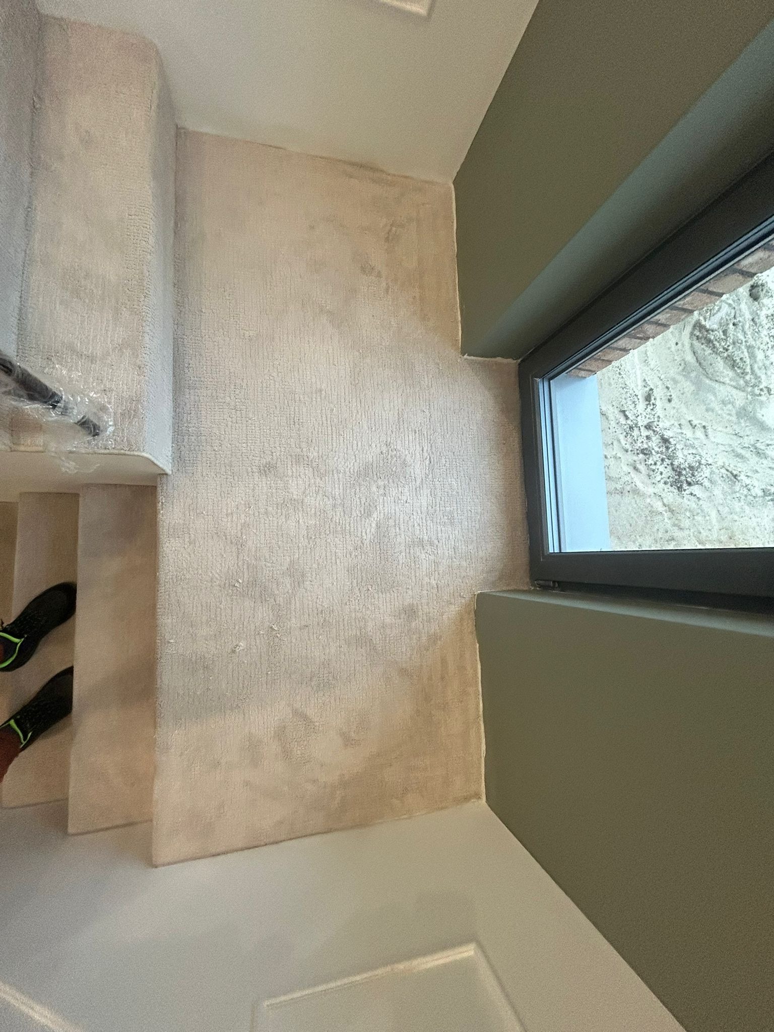 Wykończone schody z beżową wykładziną dywanową, widok z góry. Obok ściana z oknem w ciemnej ramie i fragment podłogi. Widoczne buty na schodach.