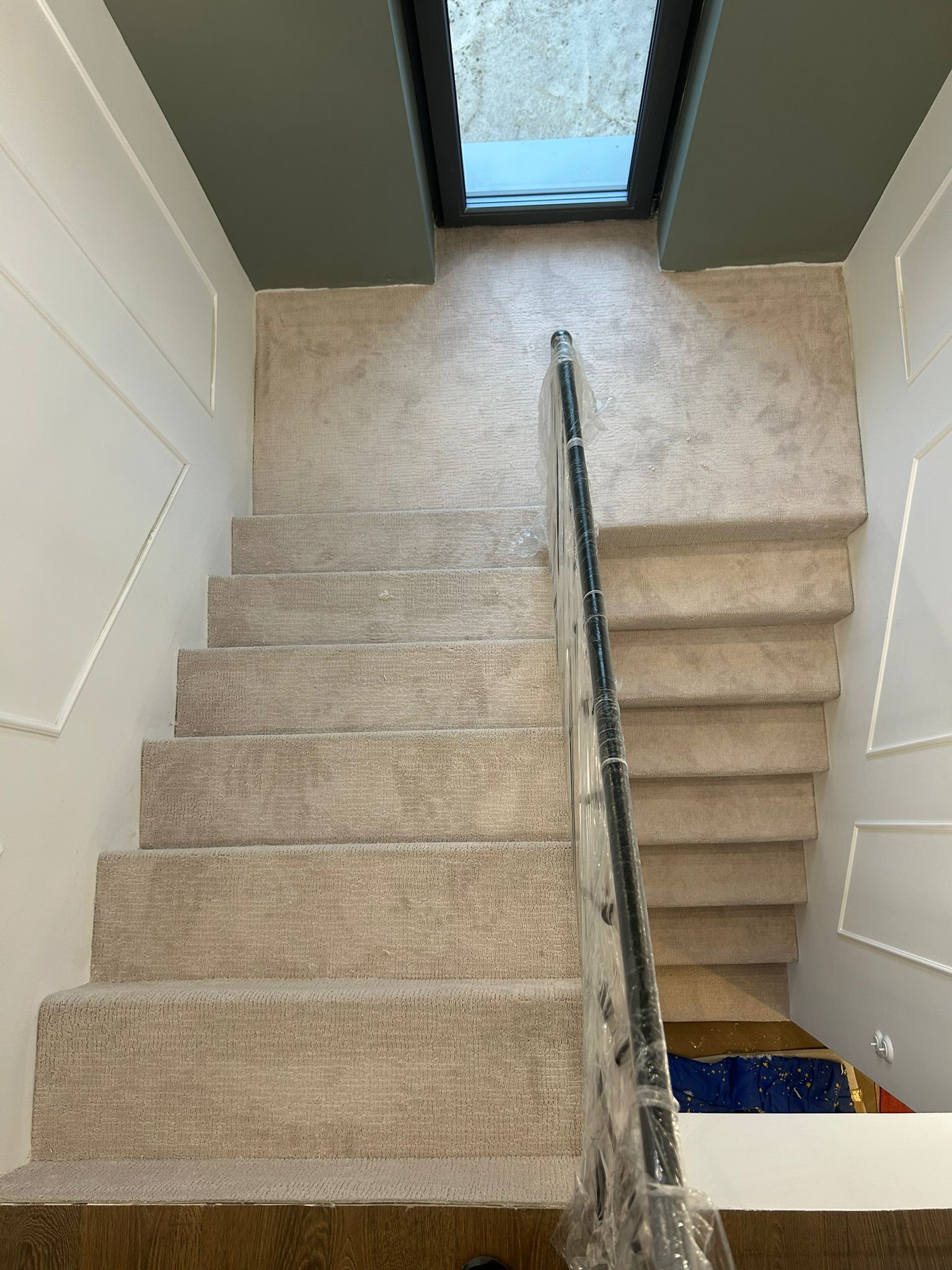 Beżowa wykładzina dywanowa na schodach z czarną poręczą, widok z góry. Wykładzina dopasowana kolorystycznie do ścian i sufitu. Okno dachowe w tle.
