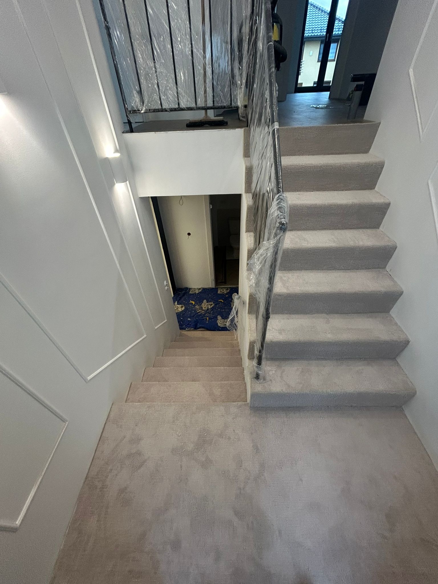 Schody wewnątrz domu wyłożone jasną wykładziną dywanową. Balustrada zabezpieczona folią ochronną. Widok z góry na dół, w tle widoczne drzwi i fragment podłogi.